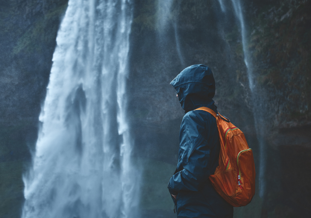 Person in dark raincoat cagoule and backpack stands next to a waterfall