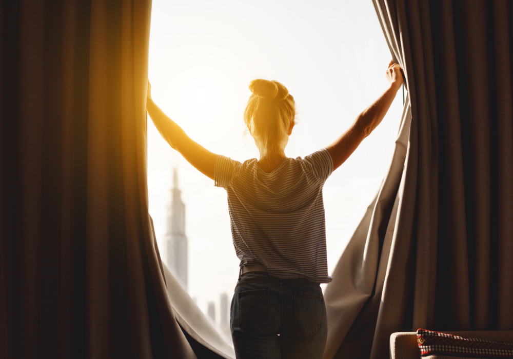 back of woman opening curtains to bright sunlight